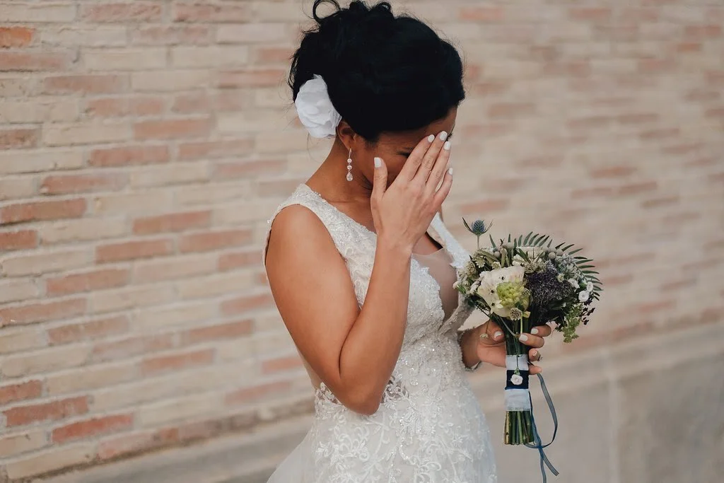 A bride in a lace wedding dress holding a bouquet of flowers while covering her face with one hand, standing against a brick wall.