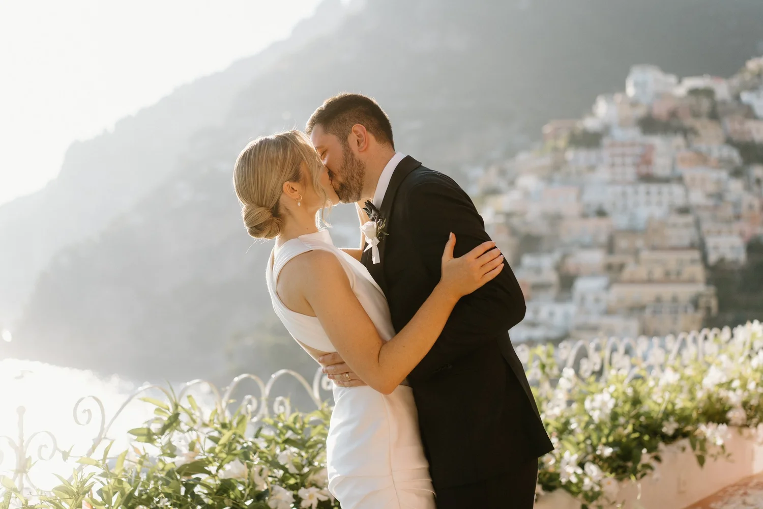 A couple dressed in wedding attire sharing a kiss outdoors with a hillside town in the background.