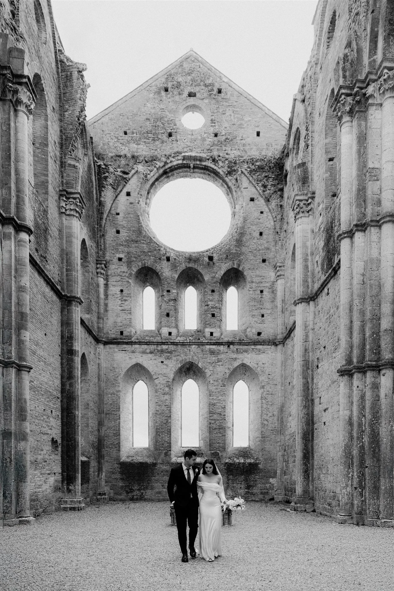 A black-and-white photograph of a bride and groom walking inside a ruined, ancient stone church with tall, empty arched windows and a large circular opening in the wall behind them.