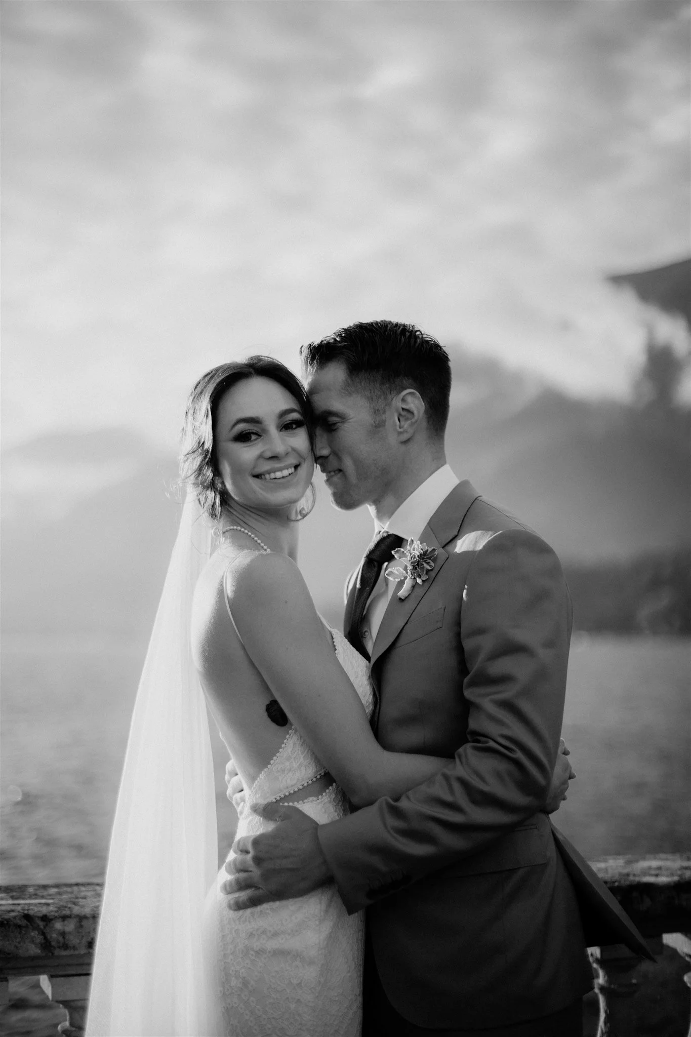 A bride and groom embracing outdoors during sunset, with the groom whispering into the bride's ear and she smiling, both dressed in wedding attire.