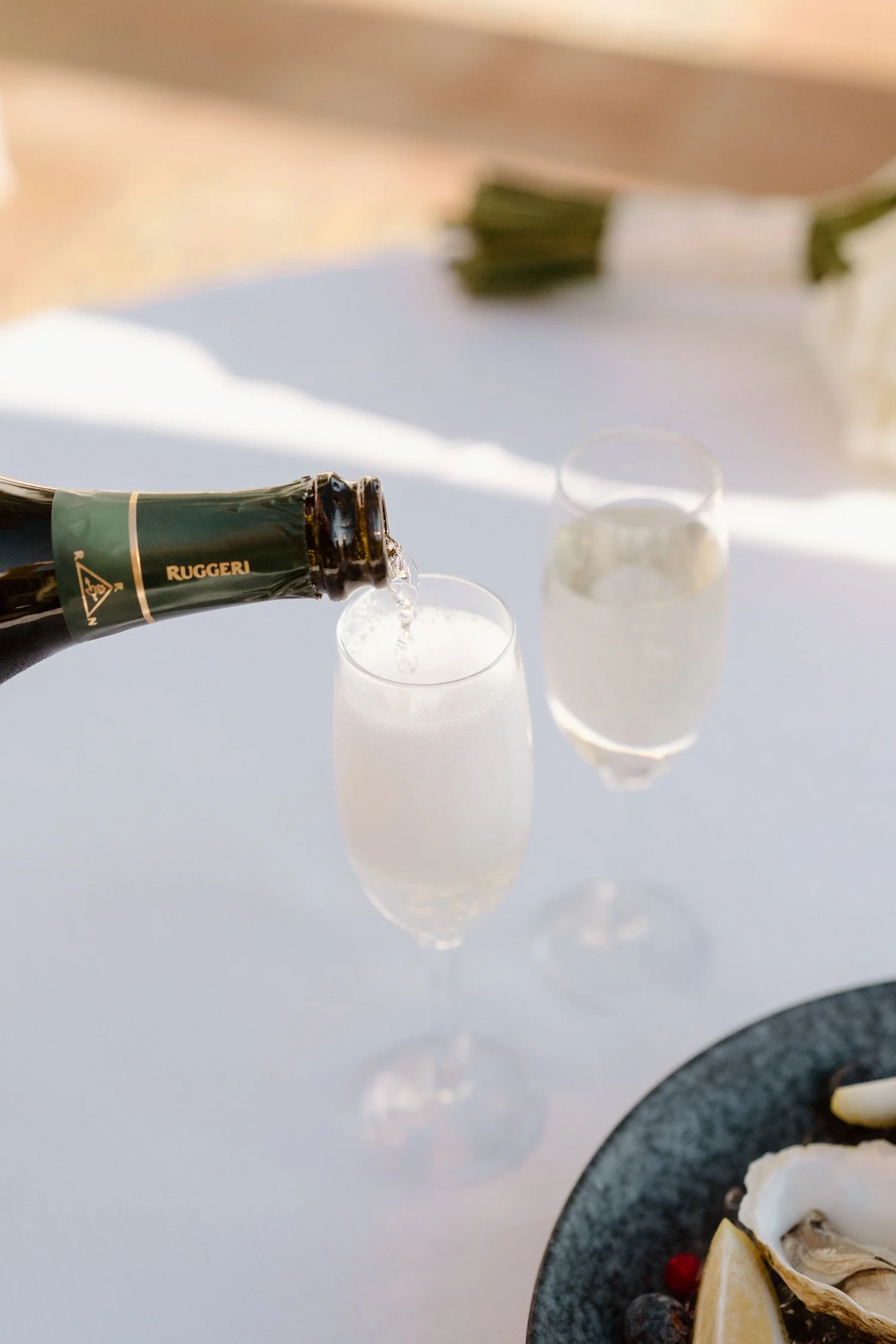 Champagne being poured into flutes on a white table, with a seafood platter featuring lemon wedges, oysters, and berries in the foreground.