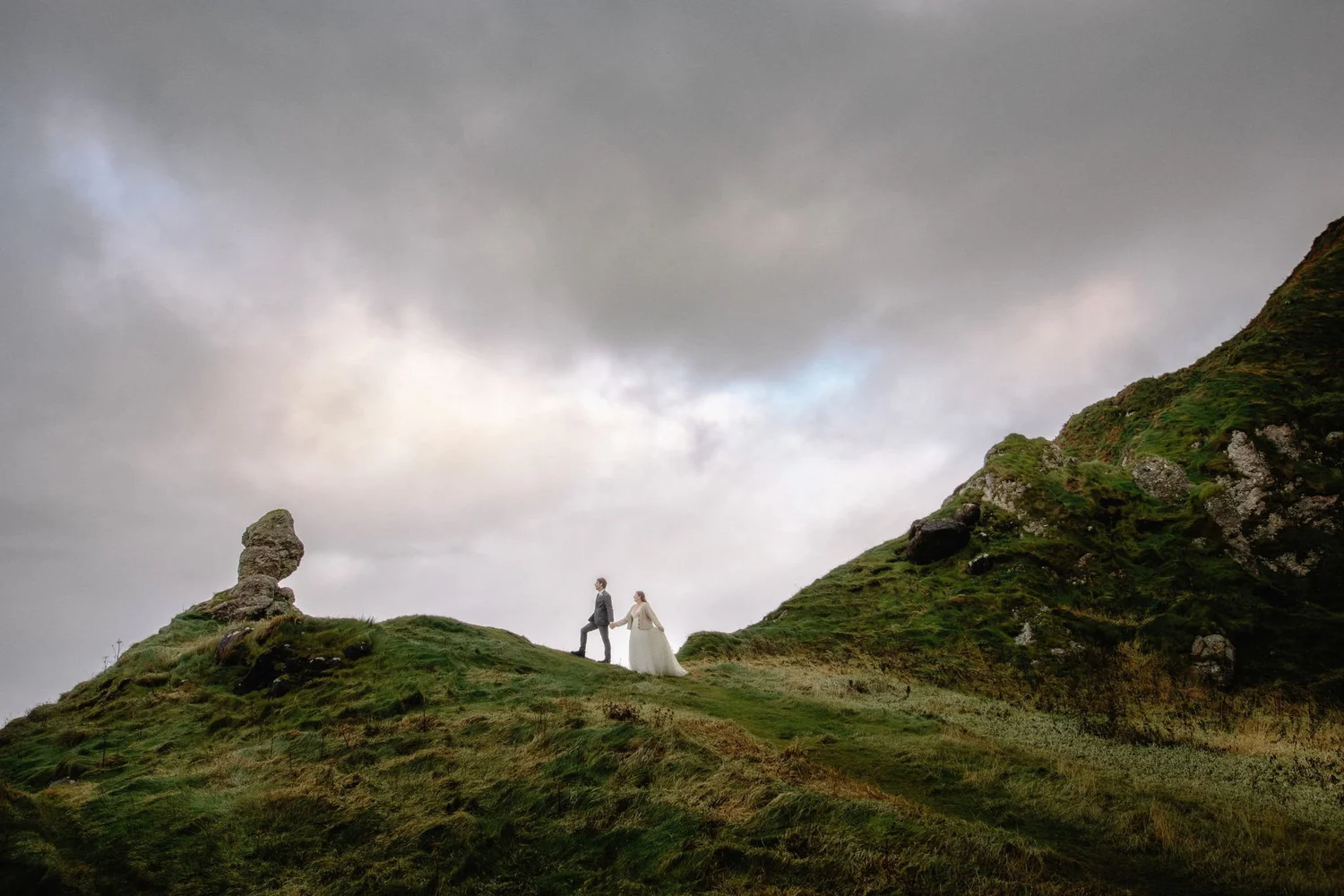 A bride and groom holding hands and walking on a grassy hill with rocky terrain and green vegetation, under a cloudy sky.