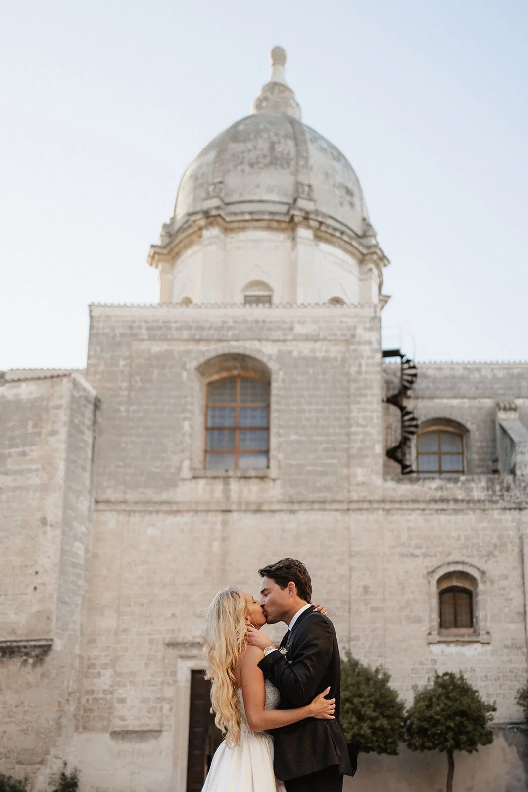 A couple kissing in front of a historic stone building with a large domed tower, during sunset or twilight.