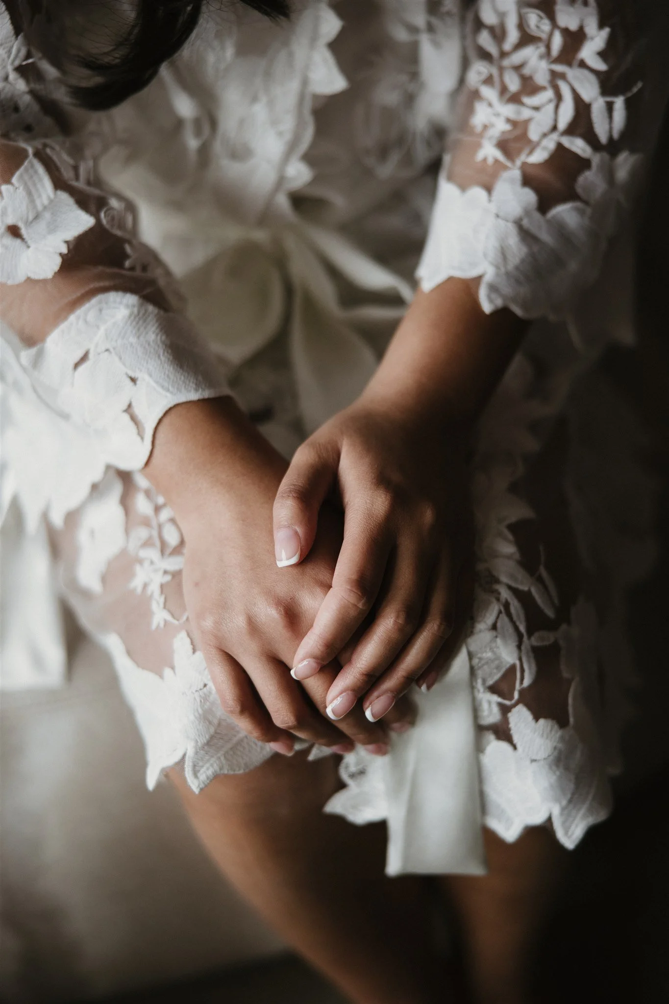 Close-up of two hands holding each other, wearing a white lace dress with intricate floral patterns.