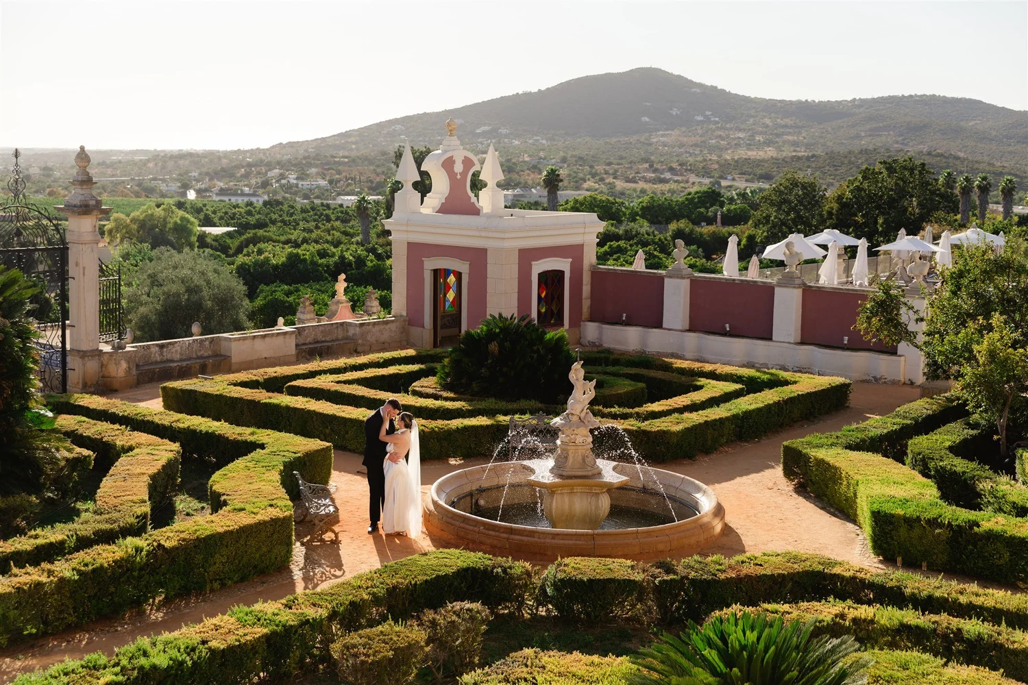 Couple in wedding attire sharing a moment in a landscaped garden with fountain, trees, and a castle-like building with stained glass windows, mountain background.