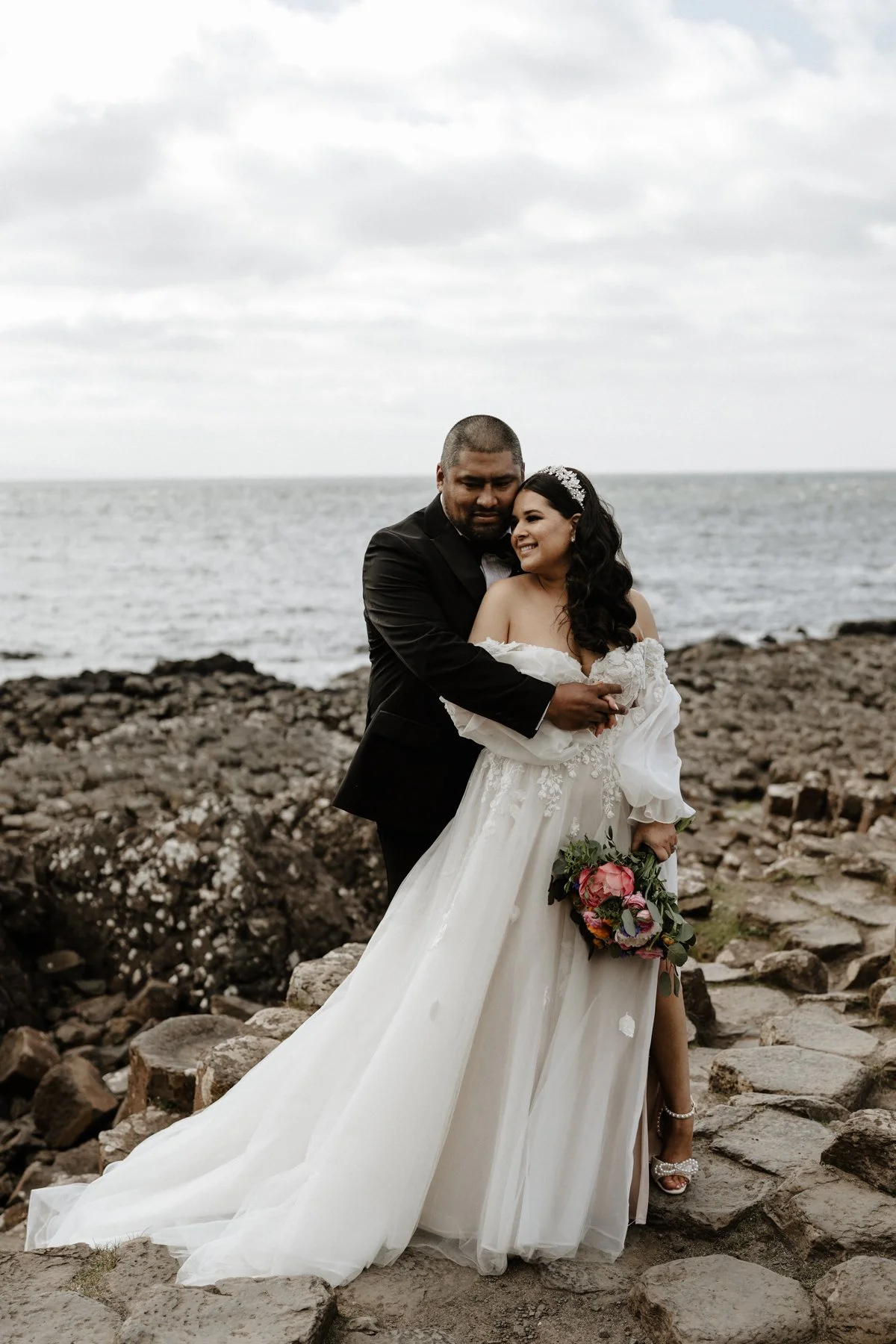 A bride and groom standing on rocky terrain by the ocean, embracing each other. The bride wears a white wedding gown with lace details and a tiara, holding a bouquet of flowers. The groom wears a black tuxedo. Overcast sky in the background.