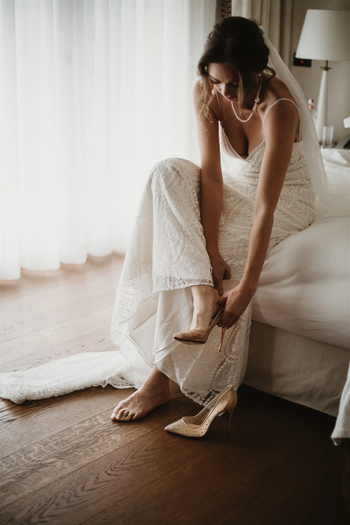A bride in a lace wedding dress seated on a bed, putting on her high heel shoes in a softly lit room.