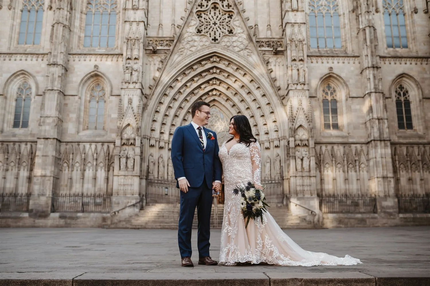 A bride and groom holding hands and smiling at each other in front of a Gothic cathedral, with detailed stonework and arched windows visible behind them.