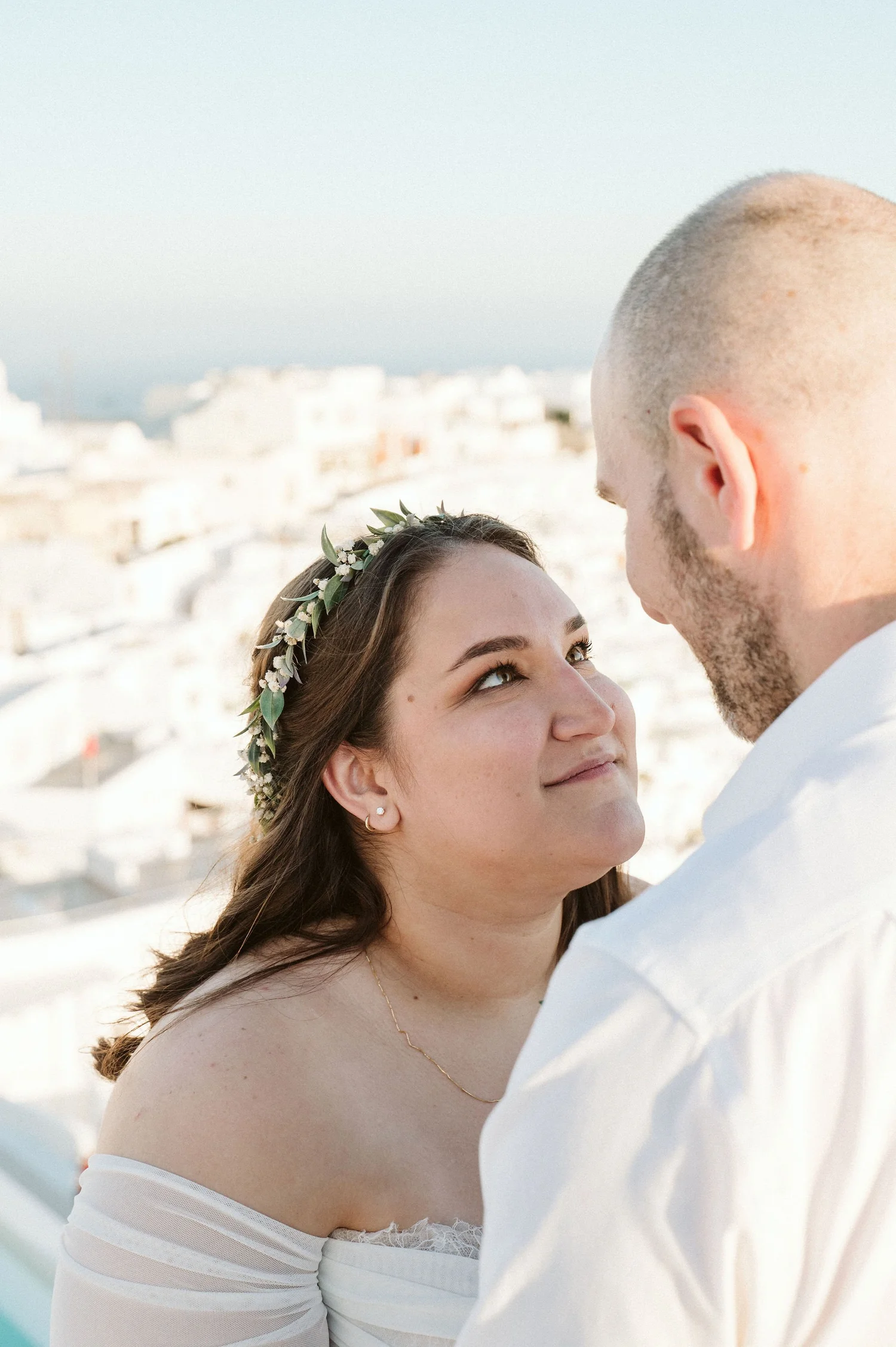A close-up of a bride and groom facing each other outdoors, with the bride wearing a floral crown and off-shoulder dress, and the groom in a white shirt, against a blurred cityscape background.