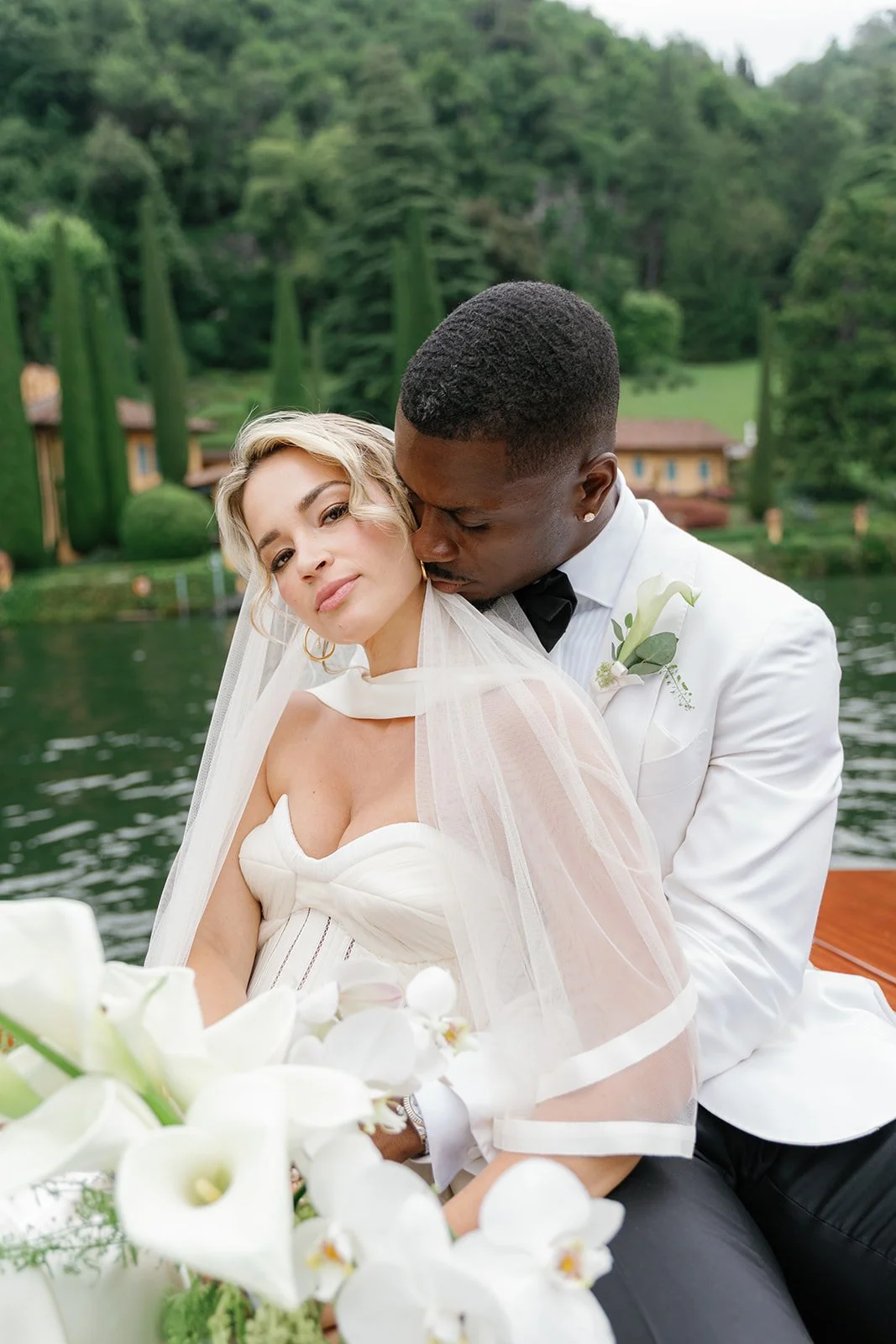 A couple dressed in wedding attire, sitting on a boat by a lake with greenery and trees in the background. The woman has blonde hair and wears a white dress with a veil, and the man has short black hair, wearing a white tuxedo with a black bow tie. They are close, with the man kissing the woman on the cheek.