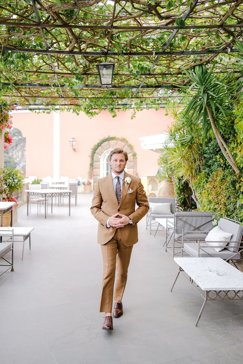 A man in a tan suit walking in a garden patio with greenery and outdoor furniture.
