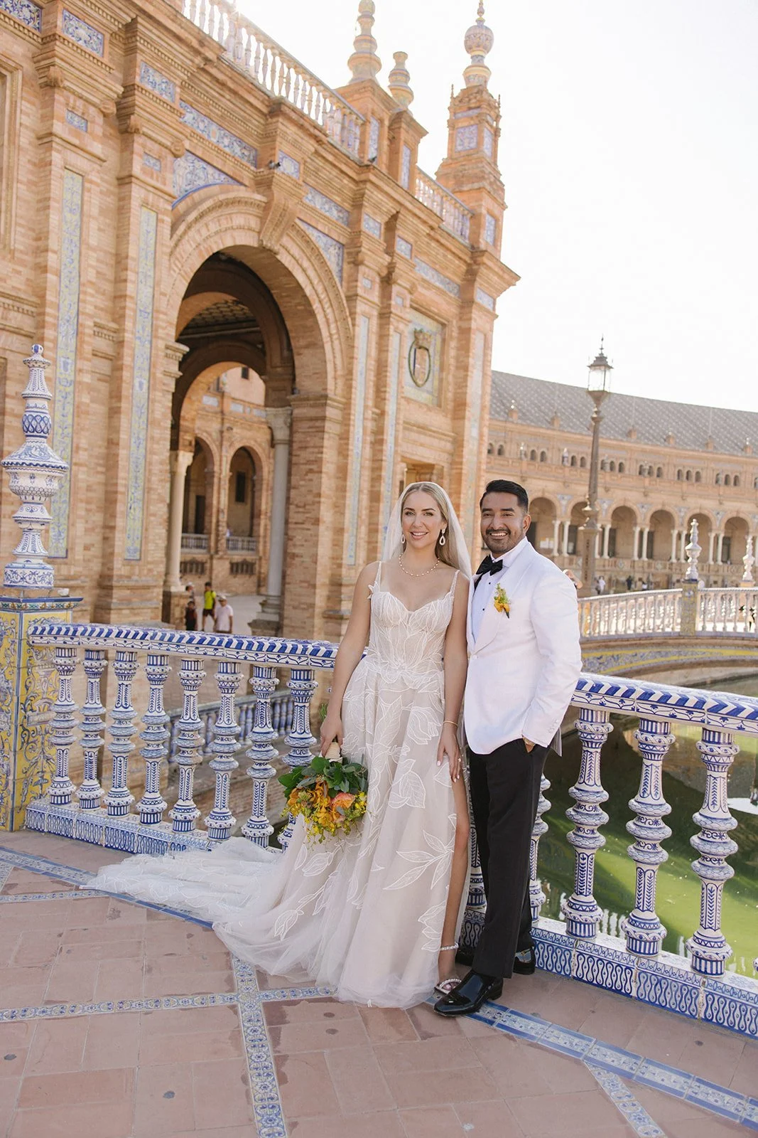 A bride and groom in wedding attire standing on a balcony with ornate blue and white tile railings, in front of a grand historic building with arched doorways and tall spires, during a wedding photo shoot.