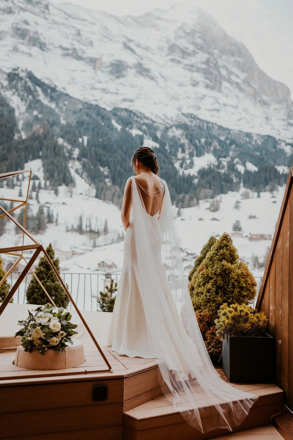 A woman in a white dress standing on a wooden patio, looking out at snow-covered mountains and trees, with potted shrubs and a bouquet of white roses nearby.