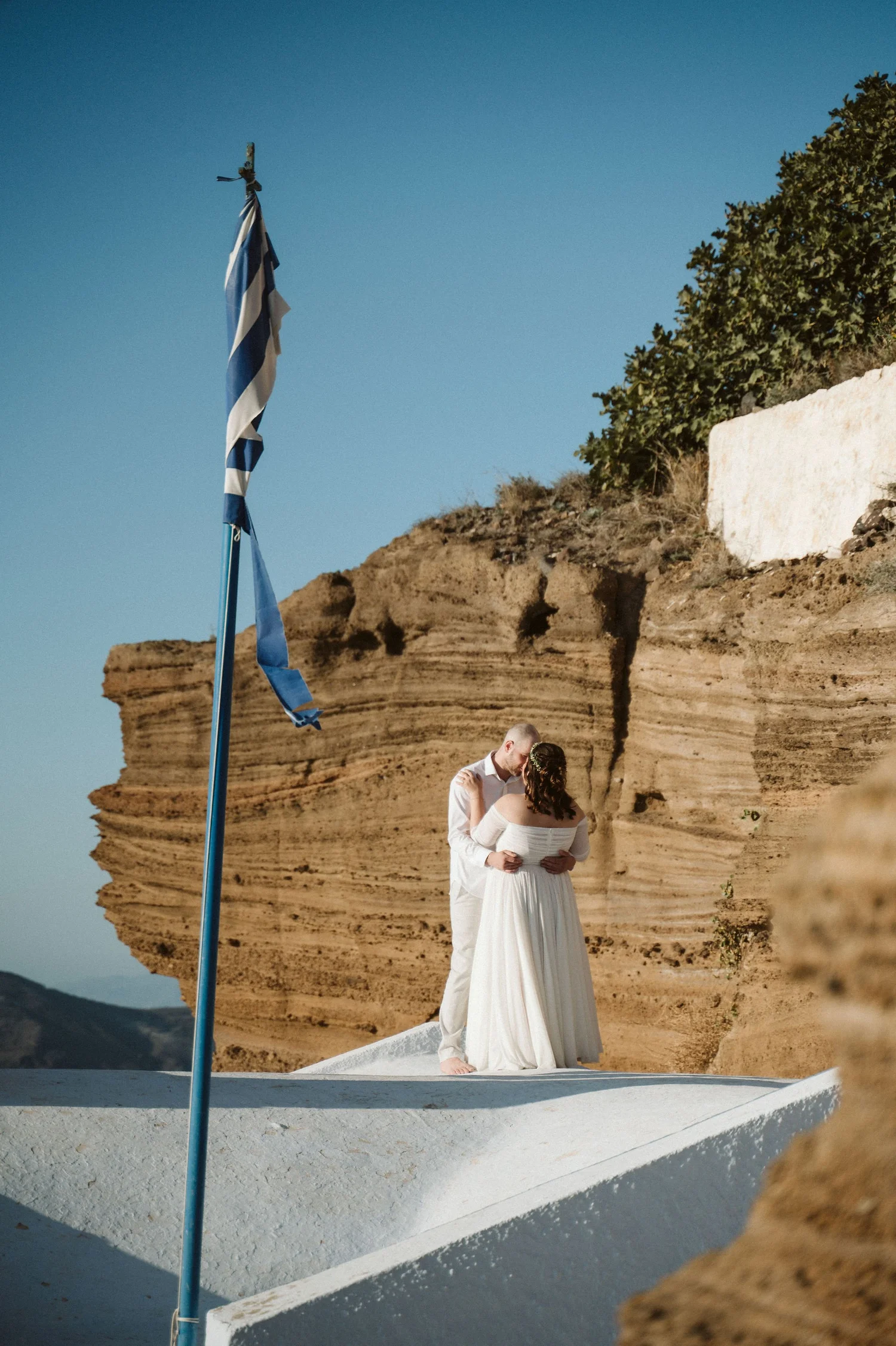 A bride and groom standing together on a white platform against a rocky cliff and a clear blue sky, with a blue and white striped flag on a pole nearby.