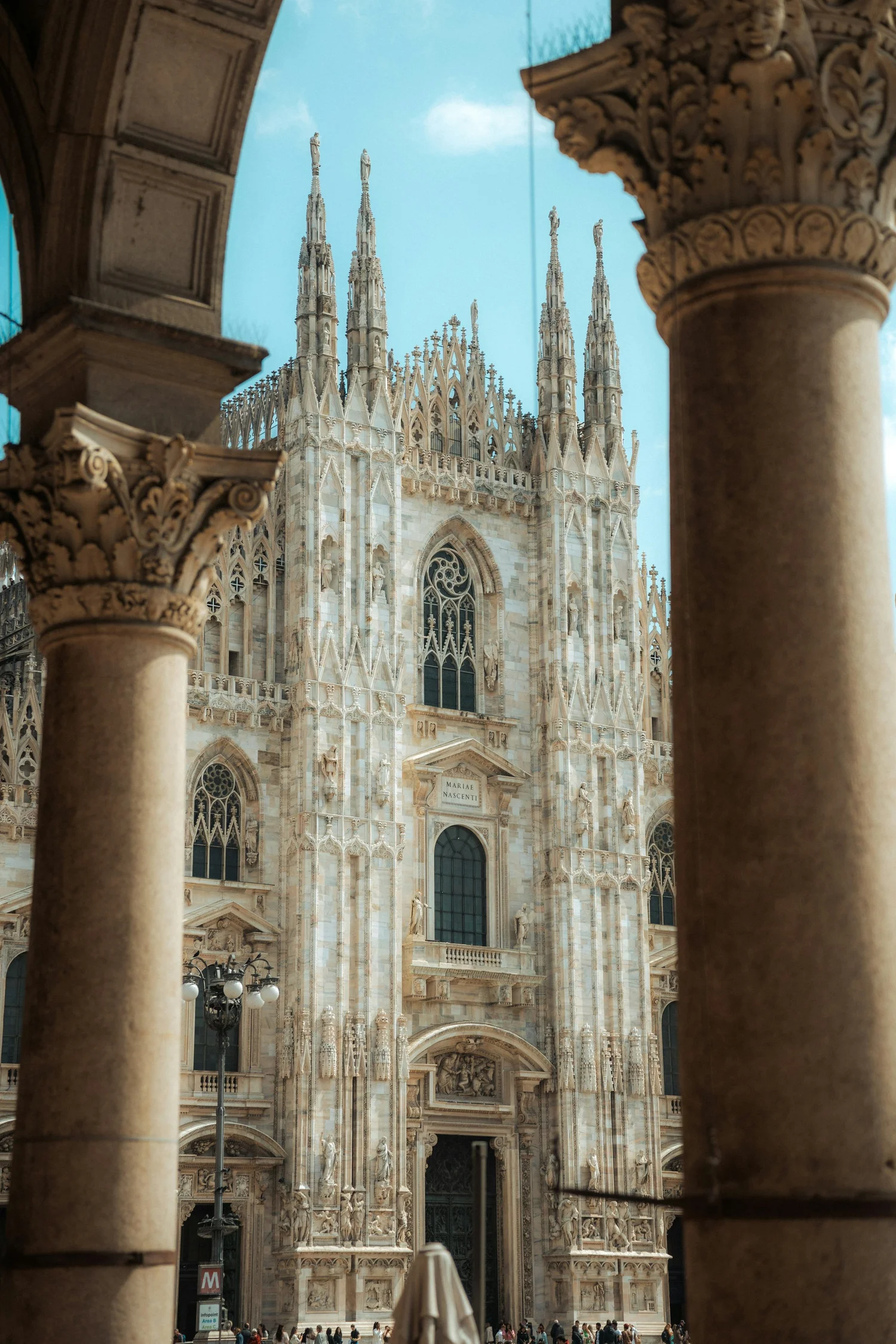 View of Milan Cathedral through stone columns and arches, showcasing detailed Gothic architecture with statues and spires against a blue sky.