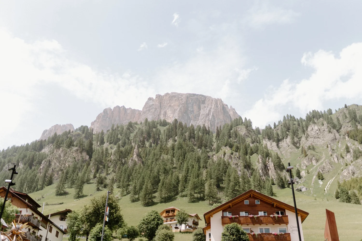 Alpine village with wooden chalets, green hillside, and rocky mountain in the background under a cloudy sky.