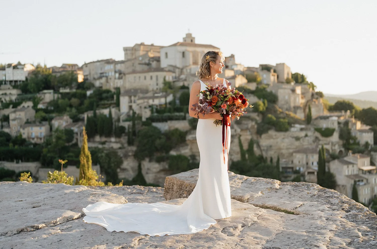 A bride holding a bouquet of flowers on a rocky ledge with a hillside town in the background.