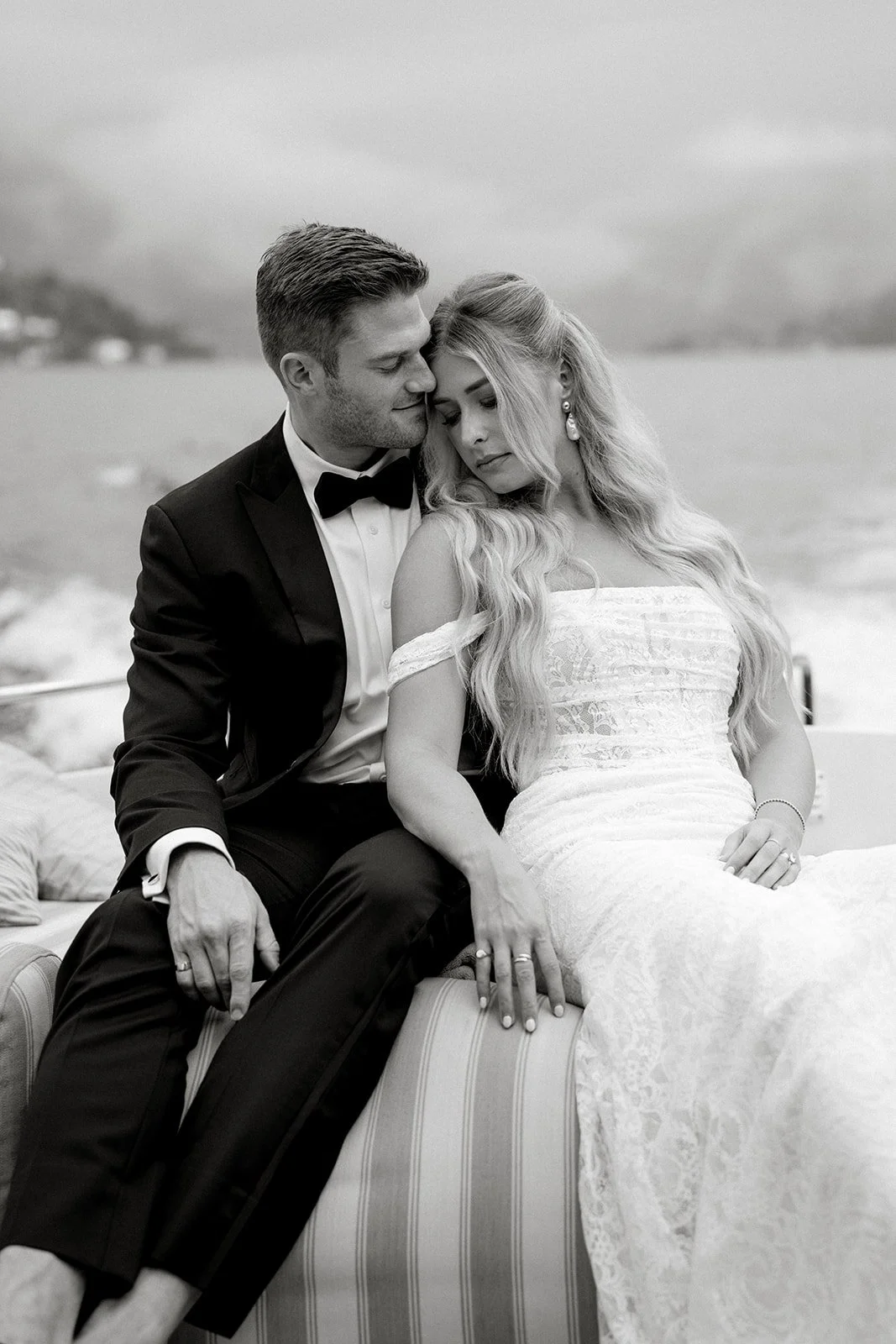 Black and white photo of a bride and groom sitting close together outdoors by the water, with the groom in a tuxedo and the bride in a lace off-the-shoulder wedding dress, sharing an intimate moment.
