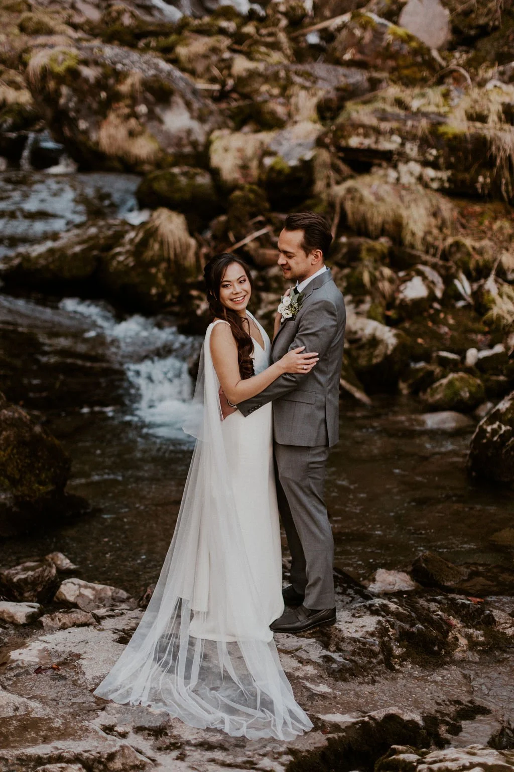 A newlywed couple standing on rocks beside a flowing creek, embracing and smiling at each other with a rocky, moss-covered hillside in the background.