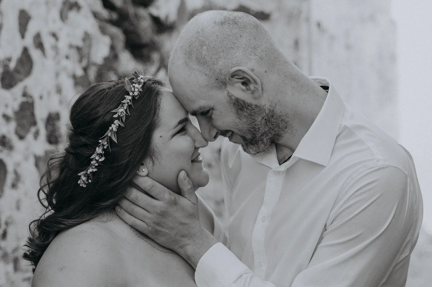 A black and white photo of a couple, with their foreheads touching, smiling and holding each other's faces. The woman has a floral headband and long dark hair, while the man has a shaved head and a beard. They are outdoors against a stone wall.