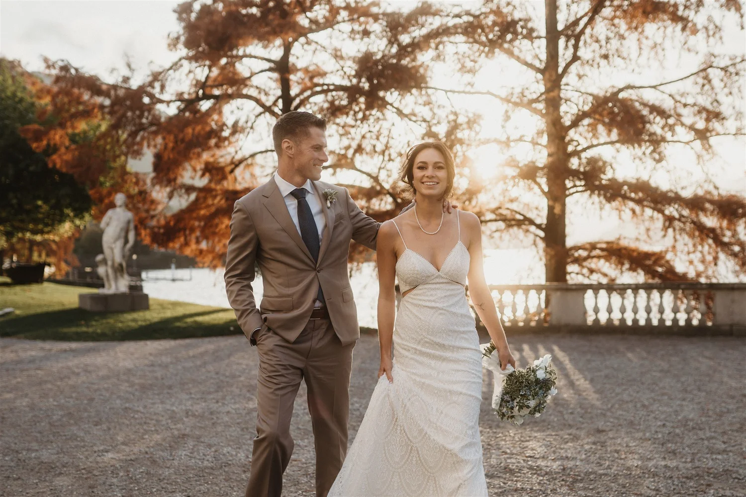 A bride and groom walking outdoors at sunset, smiling, with trees and a statue in the background.