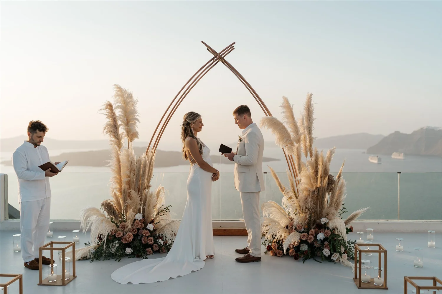 A wedding ceremony taking place outdoors with the bride and groom standing facing each other under a decorative arch made of wooden sticks and pampas grass, with a scenic view of mountains and water in the background. An officiant is reading from a book, and candles are arranged at the base of the setup.