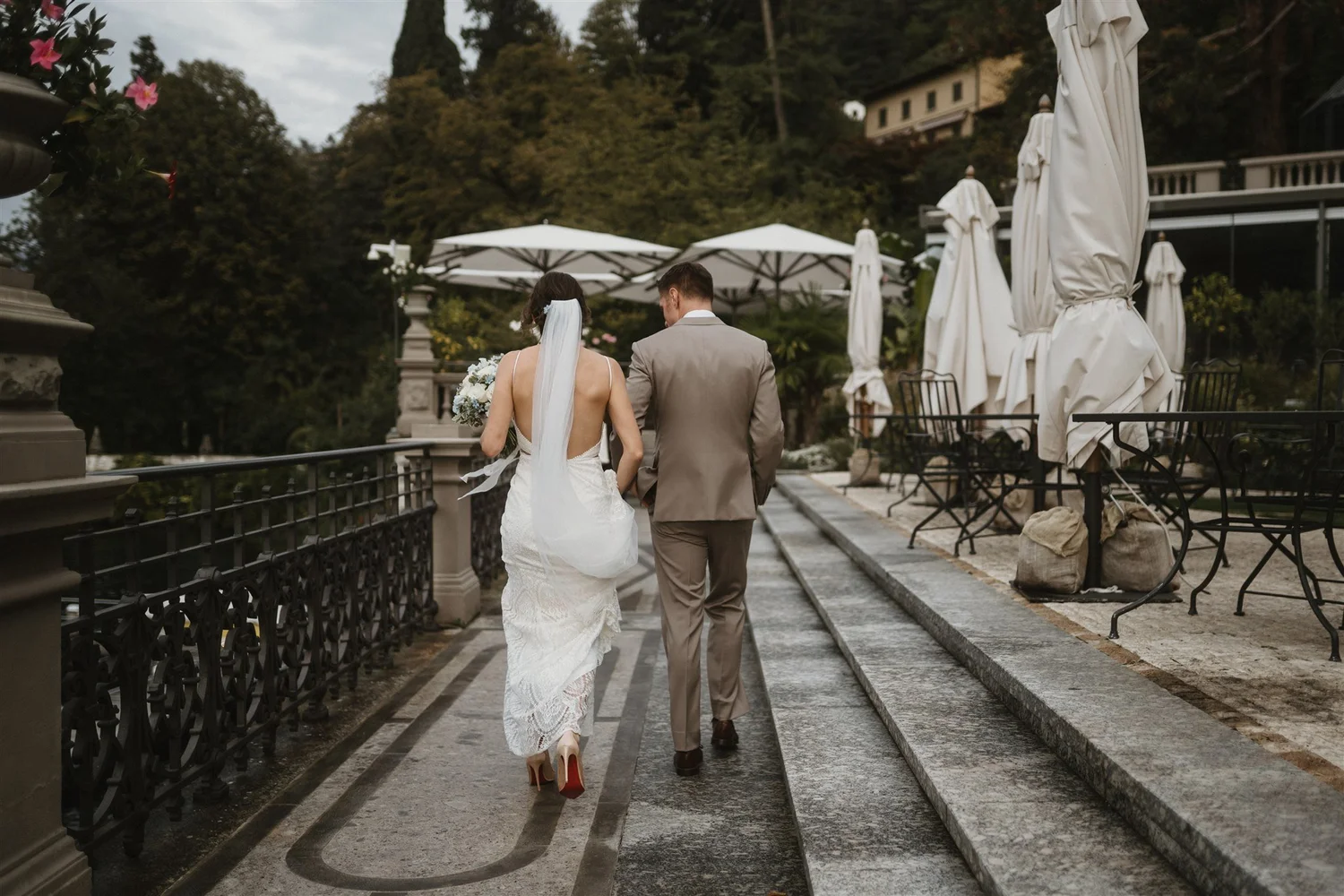 A bride and groom walking outdoors on a paved path with black metal railing, surrounded by tables with closed white umbrellas and greenery, during a wedding celebration.