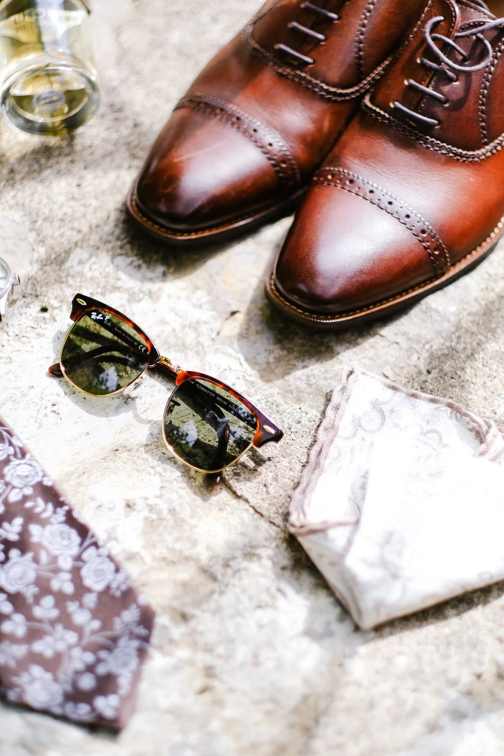 Brown leather dress shoes, sunglasses, a folded handkerchief, and a glass of whiskey on stone surface.