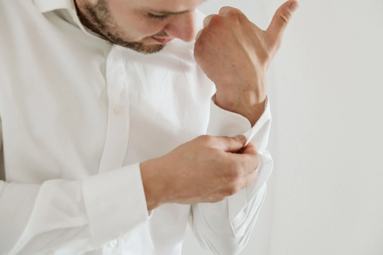 A man with a beard wearing a white shirt is adjusting his sleeve with a calm and focused expression, set against a plain white background.