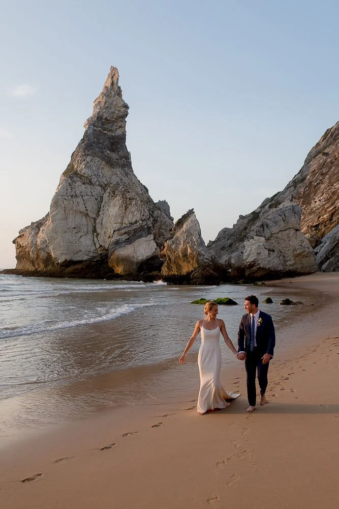 A bride and groom walking hand in hand along a sandy beach with large rock formations in the background at sunset.