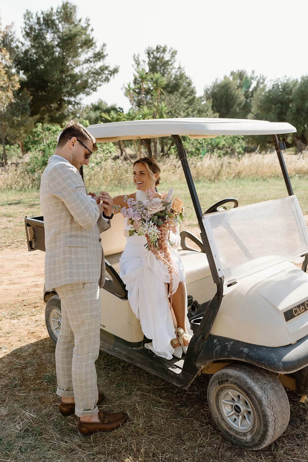 A bride sitting in a golf cart holding a large bouquet, smiling at a groom standing beside her, outdoors with trees and grass in the background.