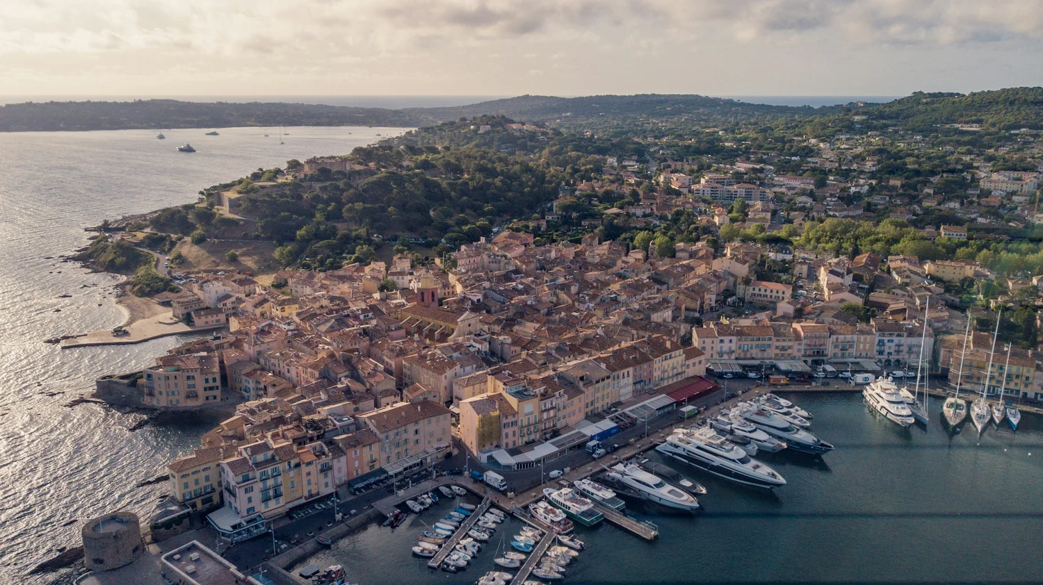 Aerial view of a coastal town with a marina filled with boats, surrounded by residential buildings and lush green hills in the background.