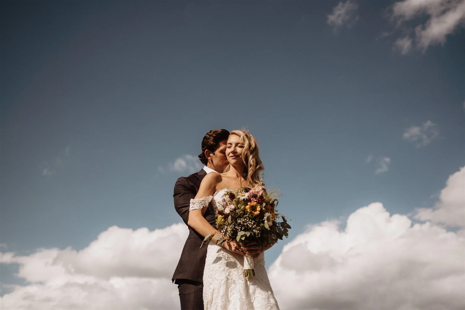 A newlywed couple standing outdoors against a backdrop of blue sky with clouds. The groom is kissing the bride's temple, and the bride is holding a bouquet of flowers, both dressed in wedding attire.