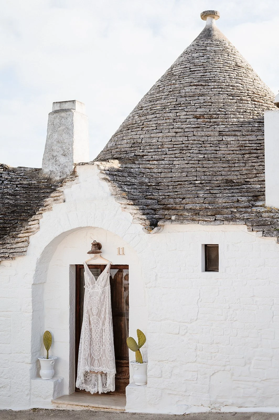 White stone house with conical roof, doorway with lace wedding dress hanging, and potted cacti outside.