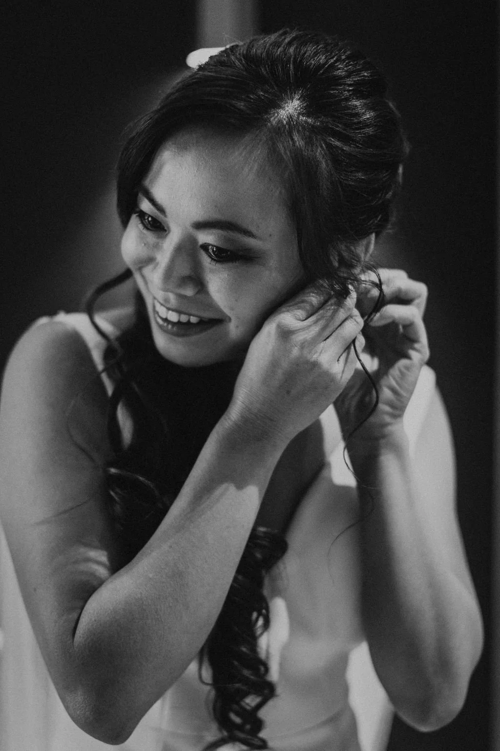 A woman with long, dark, wavy hair smiling while putting on an earring.