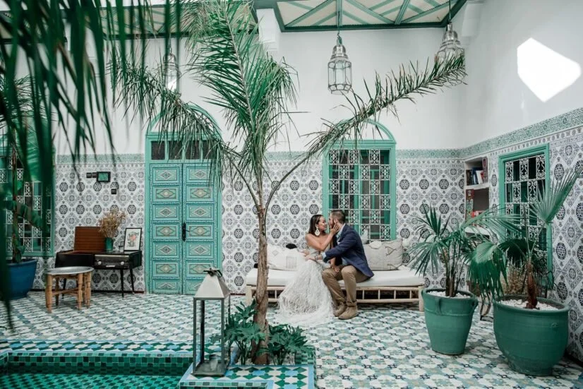 A bride and groom sitting on a white couch in a decorated indoor space with tiled walls and floor, surrounded by potted plants and large palm trees, with the bride holding the groom's face and the groom leaning in for a kiss.