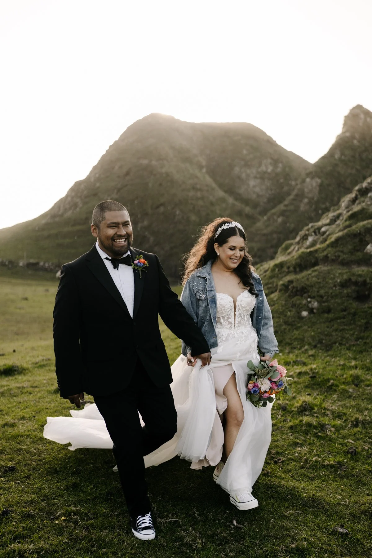 A smiling bride and groom walking hand in hand outdoors in a green field with mountains in the background, the bride holding a bouquet of colorful flowers, the groom wearing a black tuxedo with a bow tie.