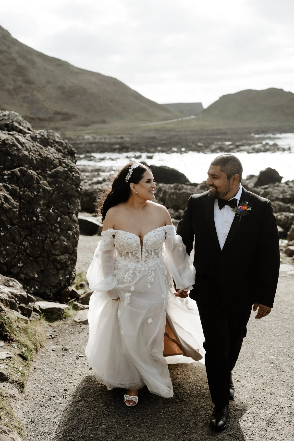 A bride and groom walking hand in hand along a rocky path by the water in a mountainous landscape. The bride is wearing an off-the-shoulder wedding dress with lace details and the groom is dressed in a black tuxedo with a boutonniere.