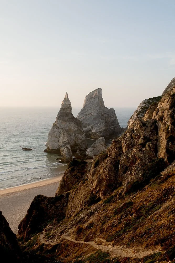 Rock formations rising from the ocean near a sandy beach, with a rugged hillside in the foreground, under a clear sky.