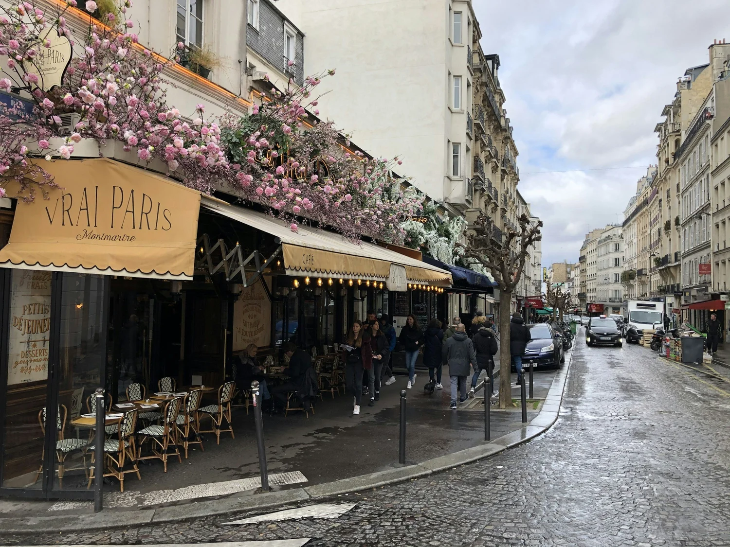 Street scene in Paris with outdoor café, pink flowers on building, pedestrians, and cars on wet cobblestone street under cloudy sky.