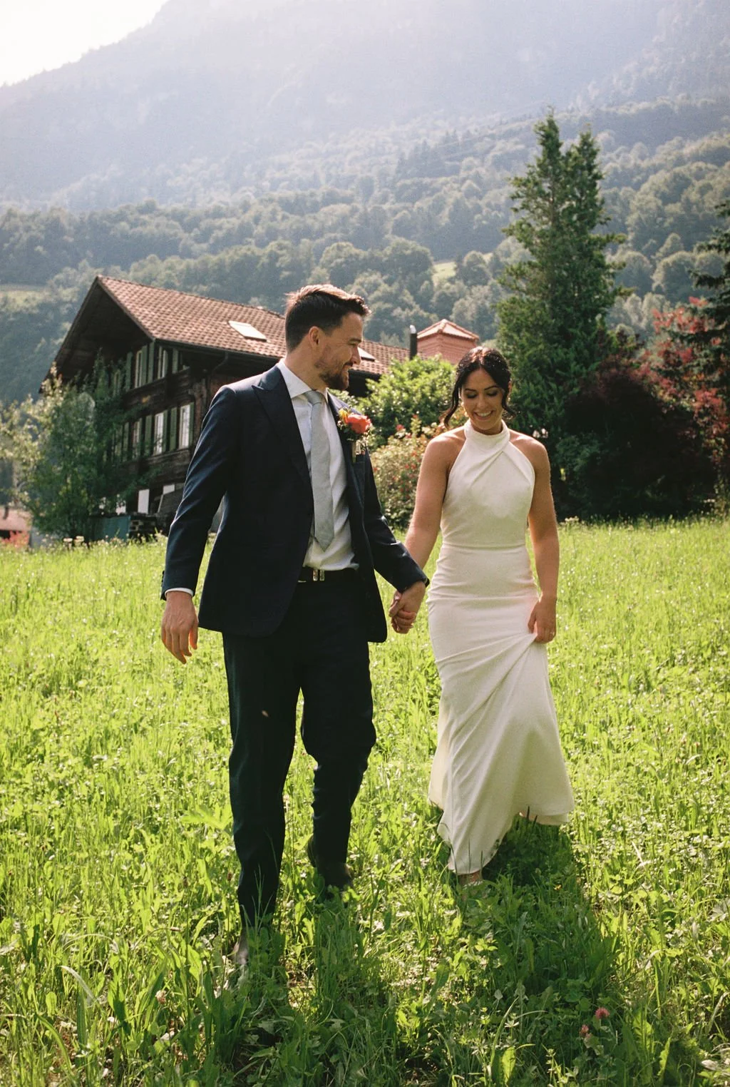 A newlywed couple walking hand-in-hand in a lush green meadow with a mountain and a house in the background, during daytime.