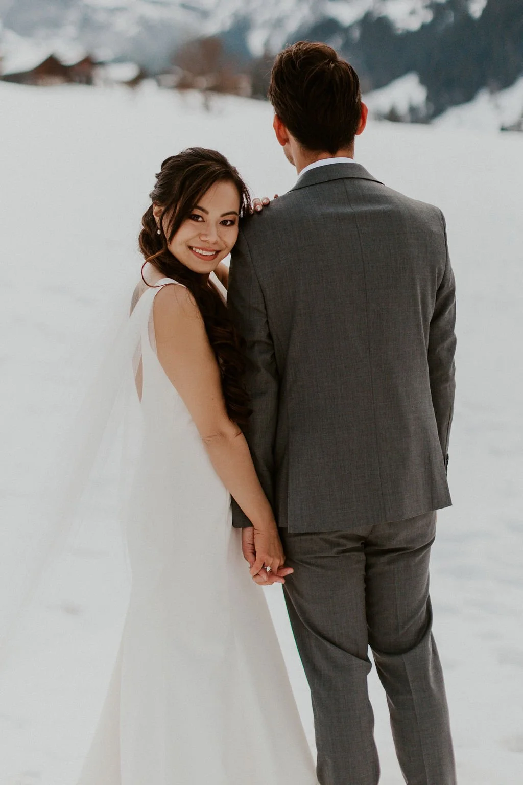 A smiling woman in a white dress holding hands with a man in a gray suit, standing outdoors in a snowy landscape with mountains in the background.