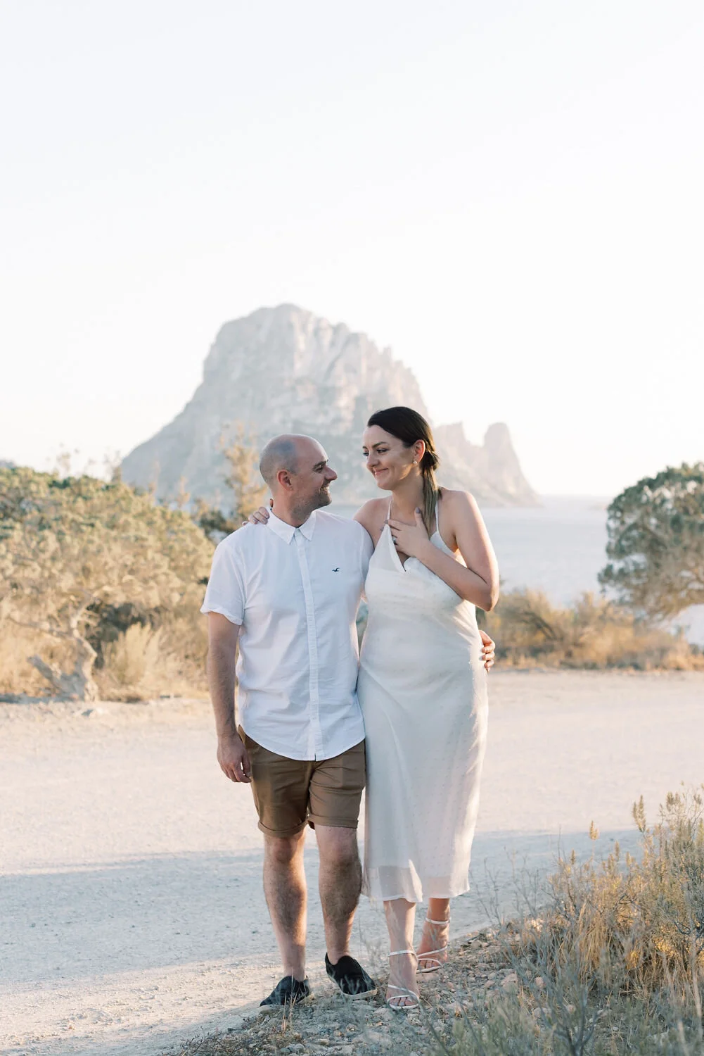 A couple walking together outdoors, holding each other, with a large rock formation and water in the background during sunset.