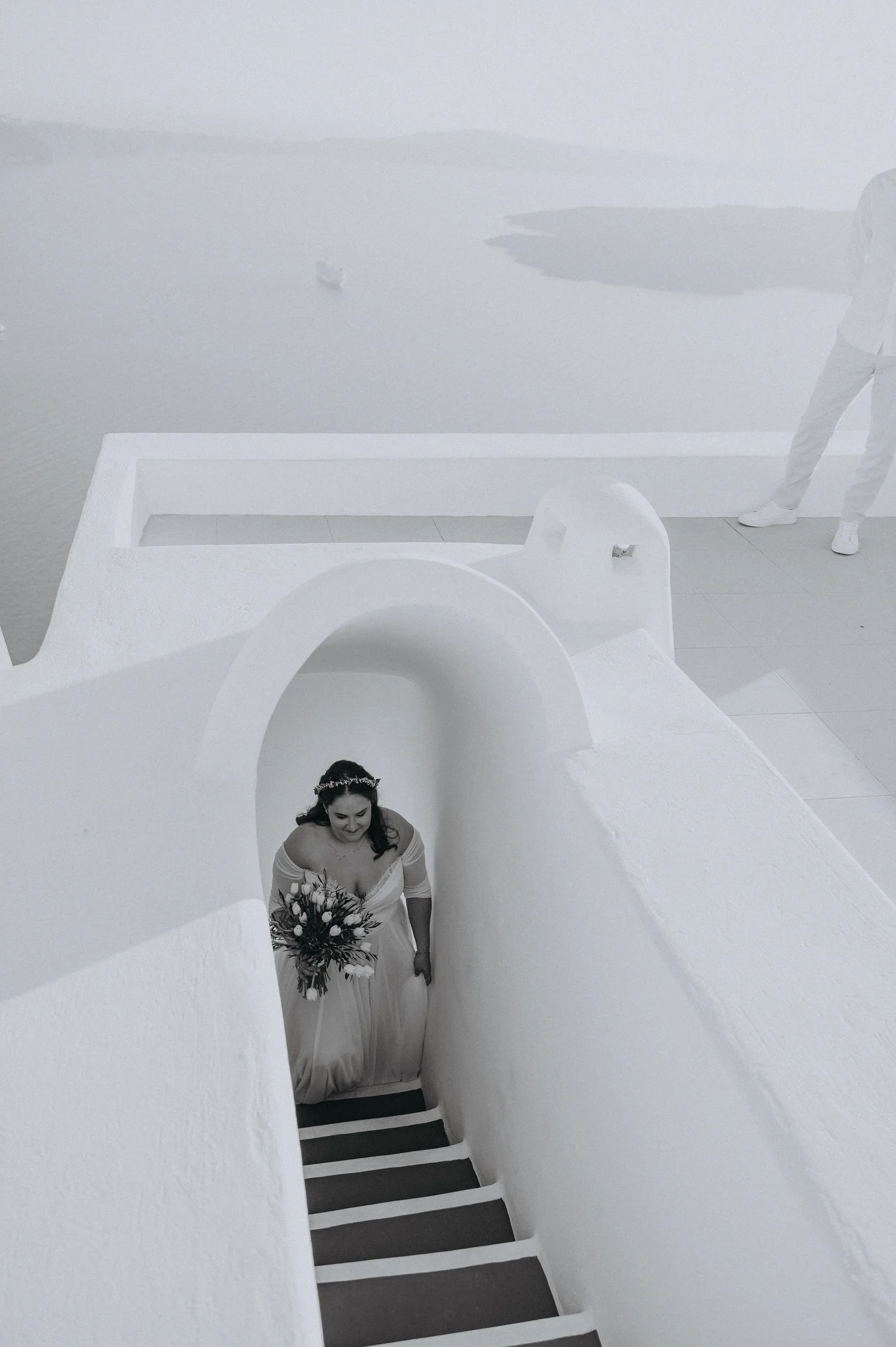 A bride with dark hair holding a bouquet of flowers walking up stairs through a white-walled archway in a minimalistic, modern setting.