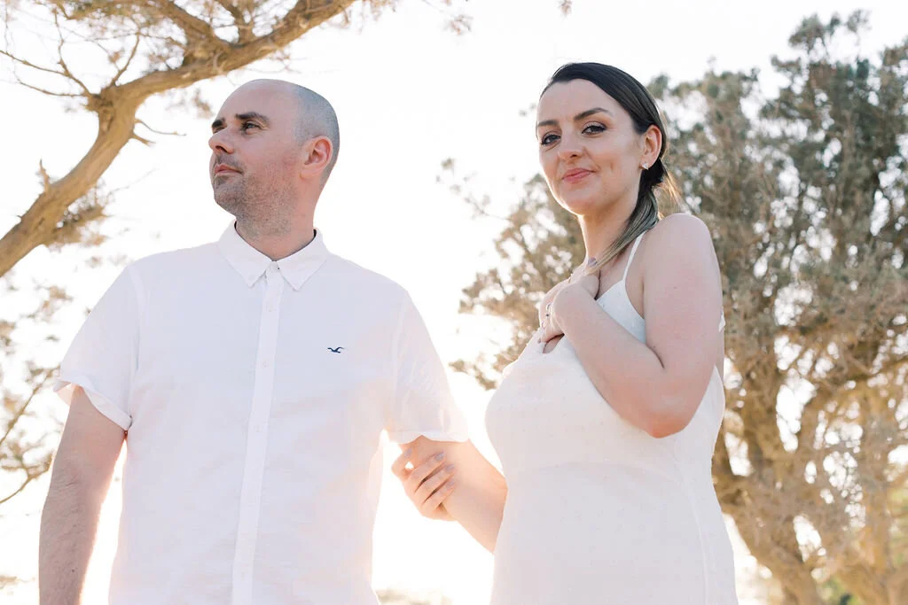A couple standing outdoors, the man dressed in a white shirt and the woman in a white dress, holding hands with trees in the background during daylight.
