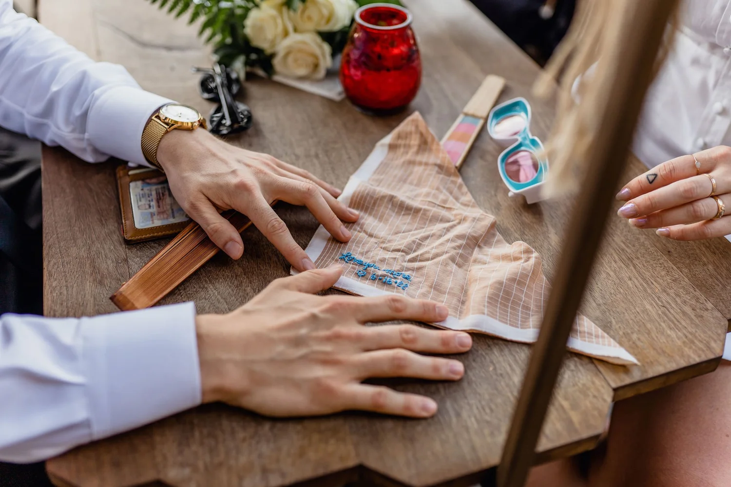 Hands of two people working on embroidery on beige fabric at a wooden table, with a bottle of water, sunglasses, and a watch visible on the table.
