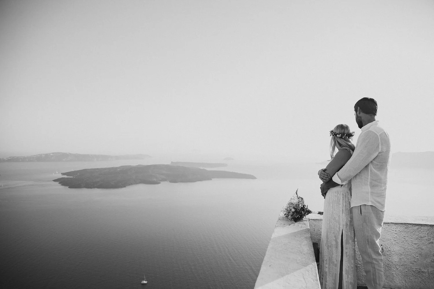A black and white photograph of a couple standing on a hill overlooking a body of water with small islands and a sailboat. The woman is wearing a floral headband and holding a bouquet, and the man is wearing a light-colored shirt and pants. They are