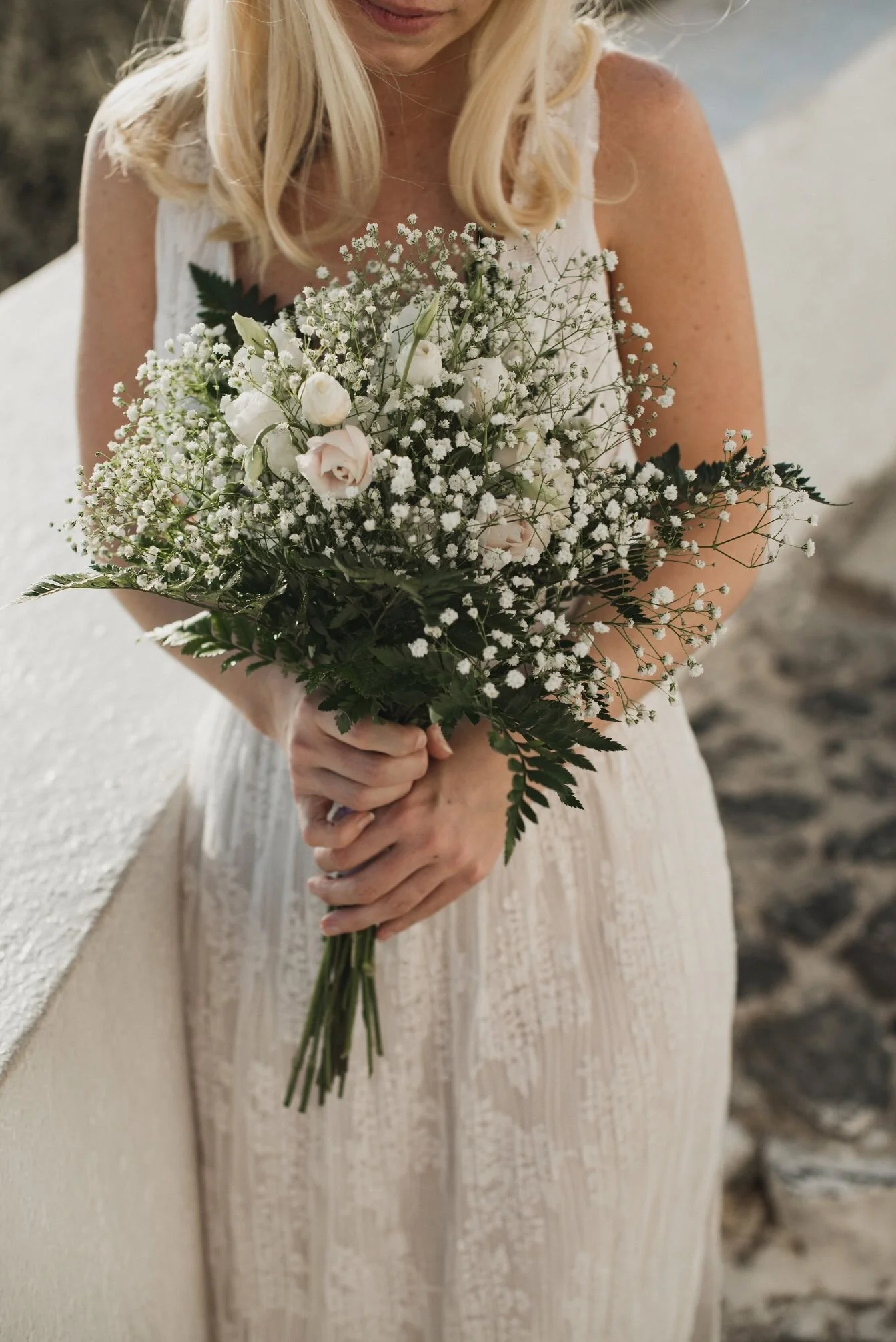 A woman in a white sleeveless dress holding a bouquet of white and light pink flowers, standing against a textured off-white wall with a blurred background.