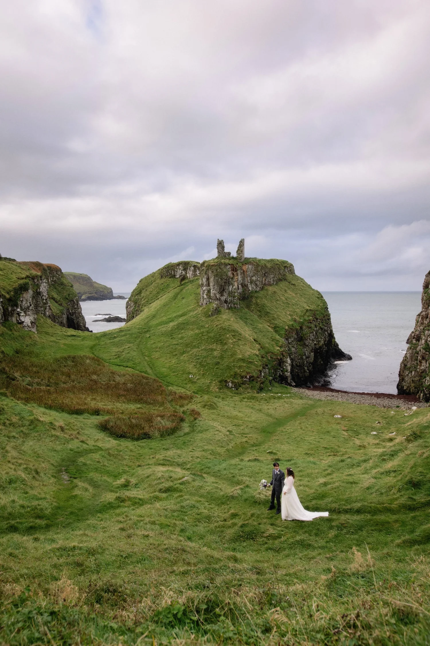 A wedding couple walking together on a grassy cliffside overlooking the ocean, with ruins on a hill in the background under a cloudy sky.