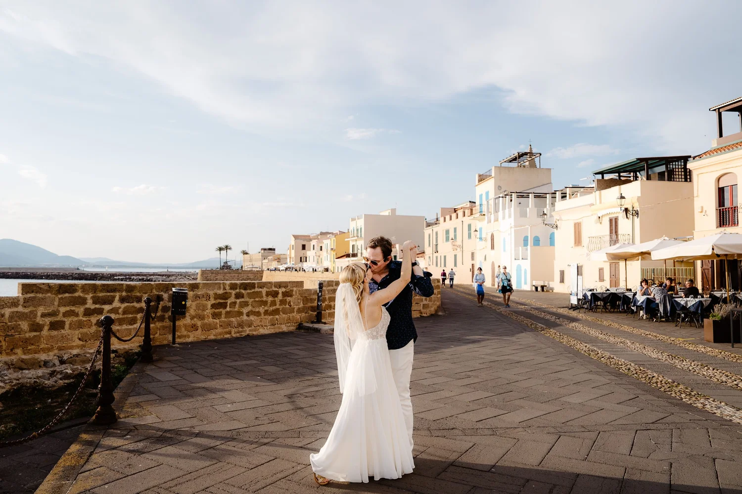 A man and a woman, dressed in wedding attire, are dancing outdoors on a paved promenade near colorful buildings by the water. The woman is wearing a white wedding dress with a veil, and the man is in a dark shirt. People are walking nearby, and there